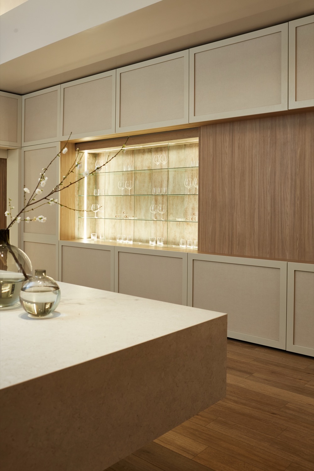 Nicho iluminado con estantes de vidrio y cristalería, integrado entre los paneles de boiserie